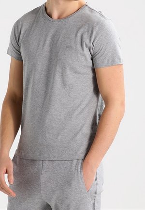 Mężczyzna w jasnoszarej koszulce z krótkim rękawem i dopasowanych spodniach dresowych, stojący na tle jednolitego jasnego tła, z jedną ręką w kieszeni.