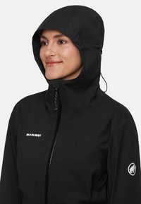 Persona che indossa una giacca a cappuccio nera Mammut con il cappuccio alzato, sorridendo leggermente, su uno sfondo chiaro e uniforme.