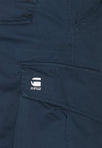 Ciemnoniebieska tkanina o gładkiej fakturze, kontrastowe szwy oraz białe logo "G-RAW" na przedniej kieszeni. Funkcjonalny design kieszeni.