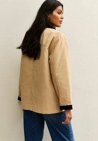 Beige canvas jack met een relaxte pasvorm en zwarte kraagdetails, gecombineerd met blauwe denim broeken, gepresenteerd tegen een neutrale achtergrond.