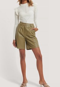 Szorty z oliwkowej sztucznej skóry z zakładkami i przednimi kieszeniami, zestawione z białą prążkowaną bluzką z długim rękawem ozdobioną falbankami.