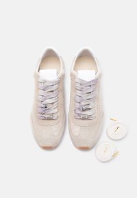 Paire de baskets en daim beige avec des lacets gris clair et des accessoires métalliques pour lacets, présentée avec deux lacets blancs supplémentaires aux embouts dorés.