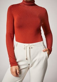 Haut rouge à col roulé côtelé associé à un pantalon blanc texturé avec cordon de serrage et accents dorés, mains posées sur les hanches.