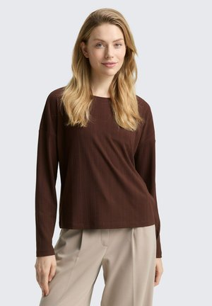Vrouw met lang blond haar, gekleed in een bruin ribbeltruitje met lange mouwen en beige broek, staand met één hand ontspannen naast haar. 