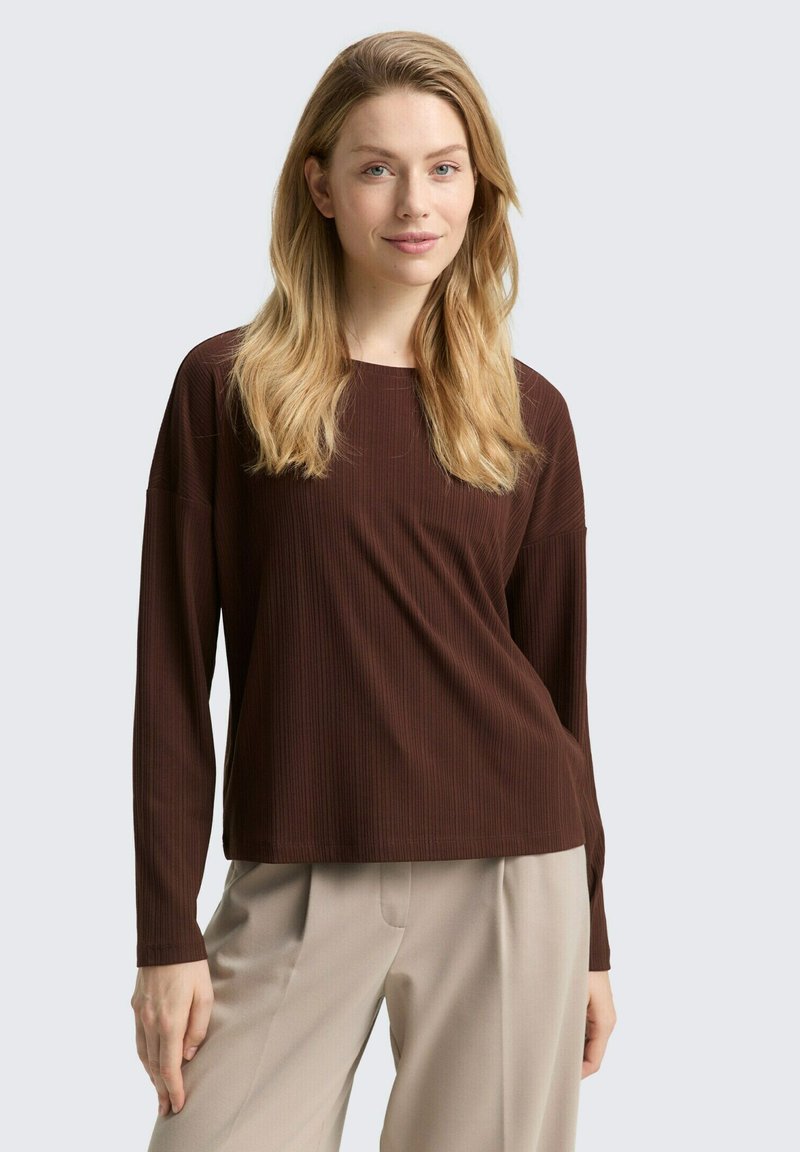 Vrouw met lang blond haar, gekleed in een bruin ribbeltruitje met lange mouwen en beige broek, staand met één hand ontspannen naast haar. 