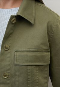 Chaqueta de algodón verde oliva con cuello abotonado, bolsillo en el pecho y una sutil textura diagonal. Presenta botones de color marrón claro y un diseño entallado.