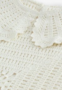 Haut en crochet crème avec un design en treillis ouvert et un col festonné. La texture douce et les détails de motif complexes ajoutent un intérêt visuel.