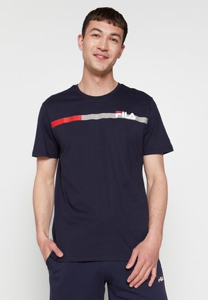 Homme portant un t-shirt et un short FILA bleu marine, debout avec les mains partiellement dans les poches, souriant devant un fond clair uni.