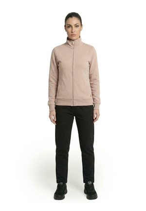 Une femme porte une veste beige zippée, un pantalon noir et des baskets noires sur un fond blanc uni.