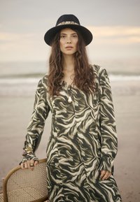 Vestido de manga larga en un patrón ondulado verde y beige, con escote en V y corte holgado. Combinado con un sombrero negro decorado con una banda.