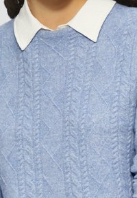 Maglione a maglia di colore azzurro chiaro con un motivo a diamante testurizzato e polsini a coste, con una camicia bianca con colletto indossata sotto.