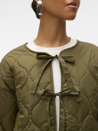 Sieviete valkā zaļu šūtu jaku ar divām priekšpusē piesietām lokām virs balta topa un lielām sudraba stīpu auskariem.