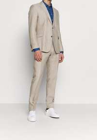 Completo in lino beige con taglio su misura, abbinato a una camicia azzurra con bottoni. Sneakers bianche completano il look. Tessuto liscio e senza motivi visibili.
