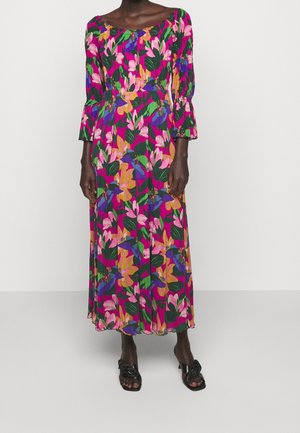 Kobieta w kolorowej, kwiatowej długiej sukni z rękawami typu 3/4 z bufkami i czarnych sandałach na obcasie, stojąca na jednolitym białym tle.