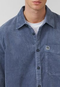 Blauwe corduroy shirt met een knoopsluiting aan de voorkant, verspreide kraag en één borstzak. Heeft een verticale ribbeltextuur en donkere knopen.