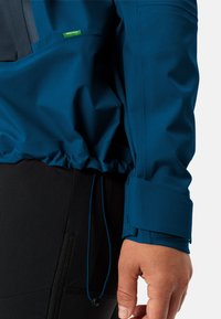 Blue pullover jacket with a textured finish, featuring adjustable cuffs and a drawstring hem. Includes a contrasting dark pocket panel.