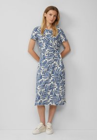 Sukienka do kolan w kolorze kremowym z niebieskimi wzorami liści, z krótkimi rękawami i wiązaniem w pasie. Połączona z beżowymi sneakersami.