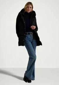 Donna che indossa un cappotto nero con cappuccio, un dolcevita nero, jeans azzurri a vita alta con una cintura e stivali neri, in piedi su uno sfondo semplice.