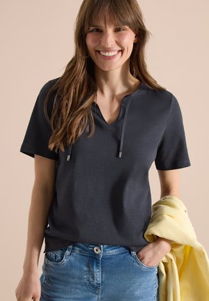Smilende kvinde med langt brunt hår iført en mørk top med korte ærmer og blå jeans, holder en foldet gul jakke i den ene hånd.