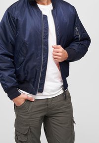 Muž v námořnickém bomberu přes bílé tričko a olivové cargo kalhoty, s jednou rukou v kapse a druhou upravující zip.