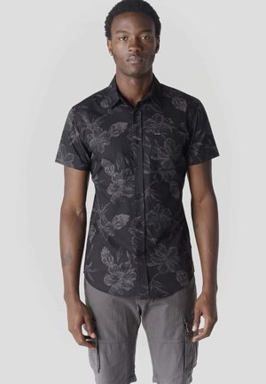 Joven de pie mirando hacia adelante, vistiendo una camisa negra de botones de manga corta con estampado floral y pantalones cortos grises estilo cargo, sobre un fondo liso.