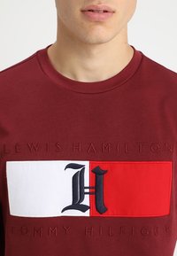 Maroon cotton t-shirt featuring a large embroidered graphic in red and white with a stylized "H" and text reading "LEWIS HAMILTON TOMMY HILFIGER."