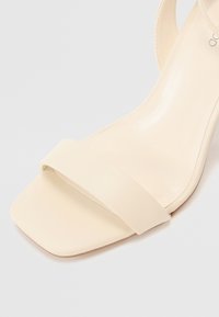 Beige høyhælte sandaler med en glatt lærfinish, firkantet tå-design og en bred reim over forsiden.