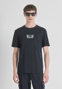 T-shirt en coton noir avec un petit graphique gris portant le texte "AM". Le modèle porte des lunettes de soleil carrées noires, associées à un pantalon cargo sombre.