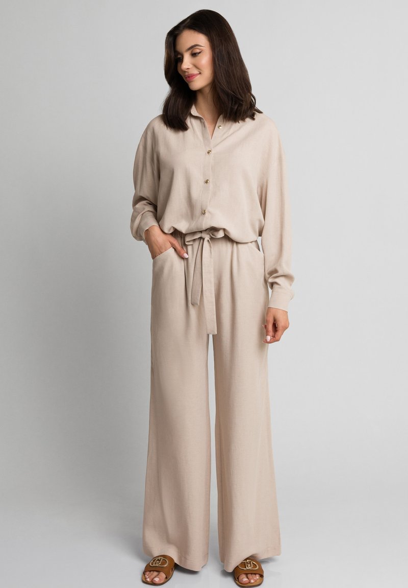 Beżowy strój z lnu, składający się z koszuli z kołnierzykiem i detalami w postaci guzików, długich rękawów oraz spodni o szerokich nogawkach z pasującym paskiem.