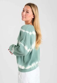 Lichtgroene gebreide trui met een losse pasvorm, voorzien van een tie-dye patroon in het wit. Ribbelstructuur en verlaagde schouders voor extra comfort.