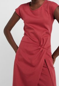 Vestido rojo hasta la rodilla con mangas cortas tipo capa, detalle fruncido en un lado y dobladillo ligeramente asimétrico hecho de tela suave y elástica.