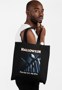 Czarna torba na ramię z graficznym wzorem przedstawiającym scenę grozy oraz tekstem: "HALLOWEEN" oraz "Sztuczka polega na tym, aby przeżyć." Gładki materiał.