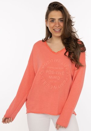 Junge Frau mit langen braunen Haaren lächelt, trägt einen lockeren korallenfarbenen Pullover mit dem Text „Positive Mind, Positive Life“ und weiße Hose.