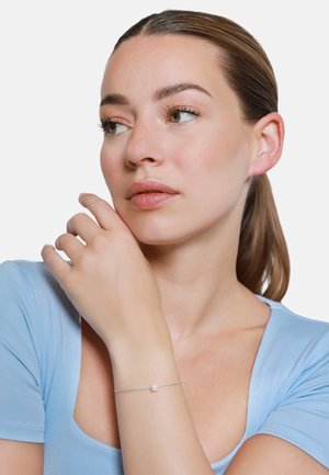 Silbernes Armband mit einer kleinen, zentralen weißen Perle, am Handgelenk getragen. Das Modell hat helle Haut und trägt ein hellblaues Oberteil.