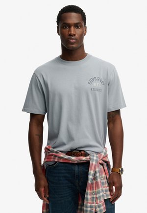 Lysegrå bomulds-T-shirt med chestprint, kombineret med blå jeans og en lyserød ternet flannel skjorte bundet om taljen.