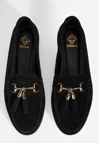 Zwarte nubuck loafers met een decoratieve gouden hardware strap met kwastjes, gestikte details en een platte zool.