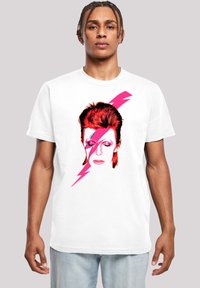 Weißes Baumwoll-T-Shirt mit einem grafischen Druck eines stilisierten Gesichts mit roten Haaren und pinken Blitzakzenten, vor einem einfarbigen Hintergrund.