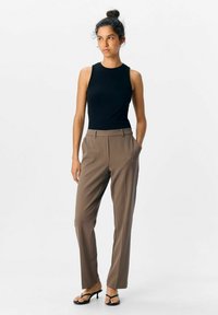 Femme debout avec les mains dans les poches, portant un haut sans manches noir, un pantalon marron ajusté et des sandales à talons noires, sur fond blanc.