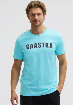 Mann mit hellbraunen Haaren, der ein himmelblaues "GAASTRA" T-Shirt und schwarze Hose vor einem einfarbigen hellen Hintergrund trägt.