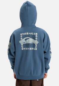 Blauwe hoodie met een groot grafisch ontwerp op de achterkant dat een golfontwerp toont, omringd door geometrische patronen in gebroken wit. Inclusief geribde manchetten.