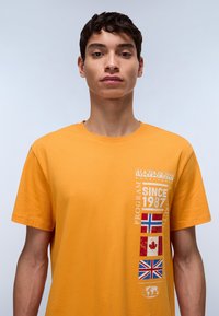 Camiseta de algodón de manga corta y cuello redondo en un brillante color naranja, con una franja vertical de banderas y texto en el lado izquierdo.