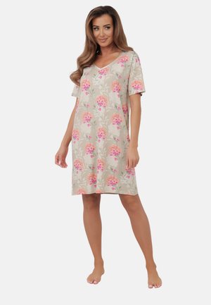 Kobieta stojąca na boso, ubrana w beżową sukienkę w kwiaty, sięgającą do kolan, z krótkimi rękawami i dekoltem w serek.