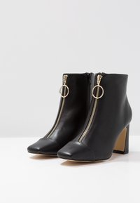 Las botas de tobillo de cuero negro presentan un diseño elegante, punta afilada y cremalleras laterales con acentos de hardware dorado. Tienen tacón y una textura suave.