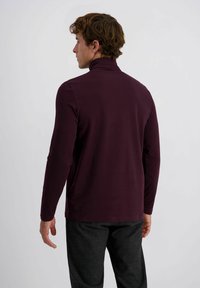 Ein Mann, der sich abwendet und einen langärmeligen dunkellila Rollkragenpullover sowie dunkle karierte Hosen trägt, vor einem schlichten hellen Hintergrund.
