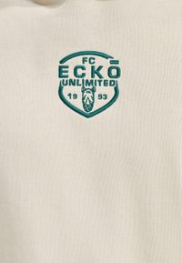 Kremfarget hettegenser med en brodert logo i teal som har en skjoldform, teksten "FC ECKŌ UNLIMITED," og året "1993."