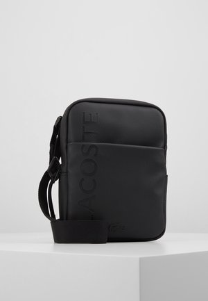 Zwarte crossbodytas met textuur, verstelbare riem, verticale Lacoste-logo en geperst krokodillenembleem op het voorvak.