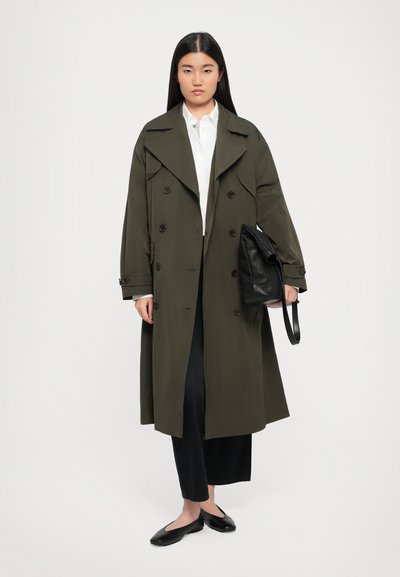 Olivgrüner Trenchcoat mit doppelter Knopfleiste, breiten Revers und langen Ärmeln. Kombiniert mit einer schwarzen Handtasche und dunklen Hosen.
