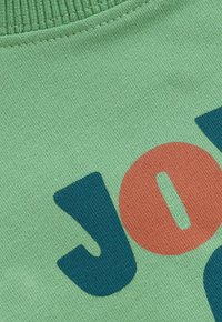 Gros plan sur un tissu tricoté vert avec un col côtelé et de grandes lettres colorées épelant "JOIE" en bleu et orange.