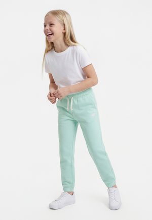 Mladé dievča sa smeje, nosí biele tričko, mäta zelené tepláky so šnúrkou a biele tenisky, stojí pred jednoduchým pozadím.
