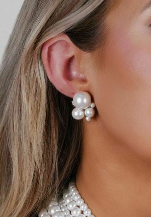 Vita pärlörhängen med flera storlekar och former, inramade i metall, uppvisade mot ljus hy och långt hår.